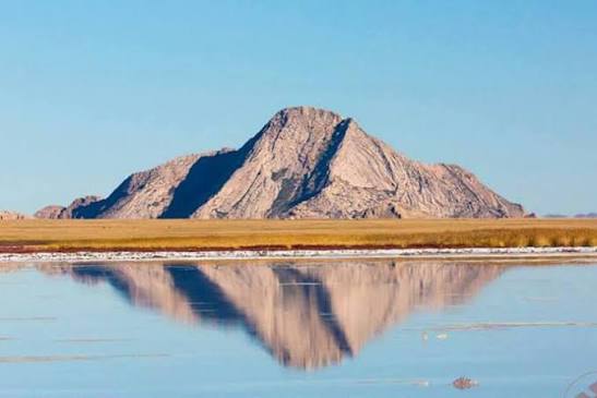 A granite mountain reflected in shallow foreground water across open steppe.